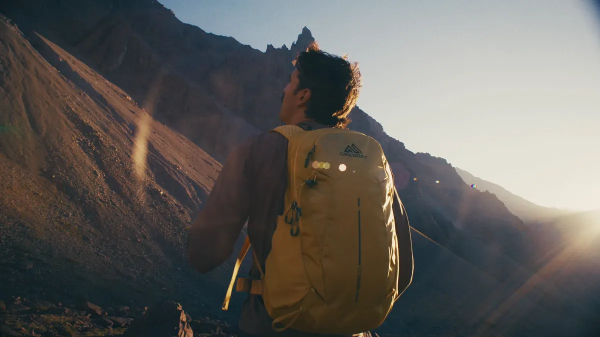 A hiker with a yellow Gregory pack looks up a mountain valley into low sun.